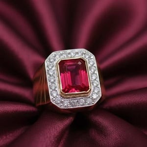 Può includere: Un anello d'oro con una grande gemma rettangolare rosso scuro, circondata da un alone di gemme più piccole e chiare. L'anello ha un design classico e audace, su un tessuto bordeaux scuro.