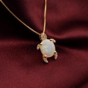May include: A gold-colored necklace with a turtle pendant. The pendant features a large, oval-shaped, opalescent stone, with small, clear gemstones on the turtle's head and flippers. The necklace is displayed on a burgundy-colored fabric.