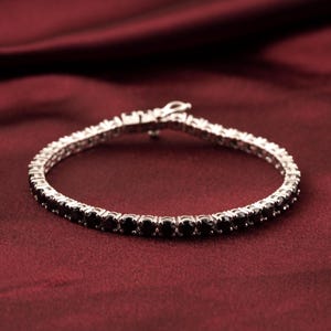 Puede incluir: Una pulsera de tenis plateada con piedras negras. La pulsera está sobre un fondo rojo oscuro. Las piedras están espaciadas uniformemente y aseguradas en un engaste plateado. El cierre es visible.