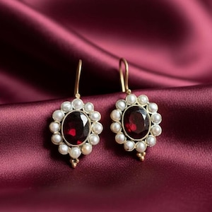 Può includere: Un paio di orecchini dorati con una gemma ovale rosso scuro al centro, circondata da piccole perle bianche. Gli orecchini sono appoggiati su un ricco tessuto di raso bordeaux, creando un'estetica lussuosa.