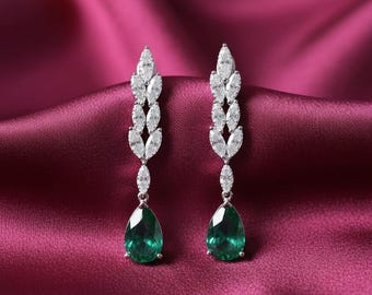 May include: A pair of elegant drop earrings. Each earring features a cluster of marquise-cut white gemstones, leading to a pear-shaped, emerald-green gemstone. The earrings are set against a deep red satin background.