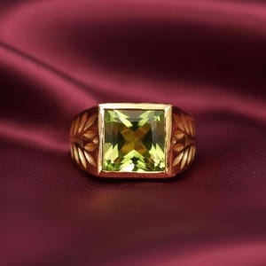 May include: A gold ring with a large, square-cut, light green gemstone. The ring has a decorative floral design on the sides and is set against a deep red background.