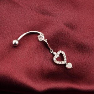 May include: A silver-colored belly button ring with a curved barbell design. The ring features a small ball on one end and a dangling heart pendant with a small leaf-shaped charm. The heart is encrusted with small, clear stones.