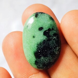 Op de afbeelding: Ovaalvormige edelsteen met een gepolijst oppervlak. De steen heeft een levendige groene basiskleur met een prominent zwart patroon. De edelsteen wordt in de hand van een persoon gehouden, wat de grootte en vorm laat zien. De steen is waarschijnlijk een decoratief item of een sieradencomponent.
