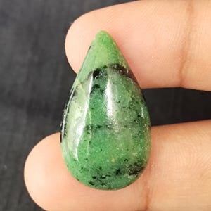 Może przedstawiać: Kształtny kamień szlachetny w kształcie łzy o wypolerowanej powierzchni. Kamień jest głównie zielony z czarnymi wtrąceniami i błyszczącym wykończeniem. Klejnot trzymany jest między dwoma palcami na ciemnym tle, ukazując jego kształt i kolor.