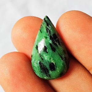 Könnte beinhalten: Ein tropfenförmiger Edelstein mit leuchtend grüner Farbe und schwarzen Einschlüssen. Die polierte Oberfläche reflektiert das Licht und zeigt die natürlichen Muster im Stein. Der Edelstein wird zwischen Fingern gehalten, um seine Größe und Form zu veranschaulichen.