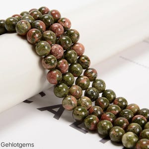 May include: A strand of round, polished beads featuring a mottled pattern of earthy olive green and dusty rose pink. The beads are uniform in size and arranged in a continuous line, showcasing natural color variations and smooth, reflective surfaces.