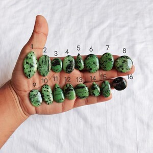 May include: A collection of sixteen green and black gemstones of various shapes and sizes, held in a hand. The stones range from oval to teardrop, with a speckled pattern. The gemstones are numbered 1 through 16.