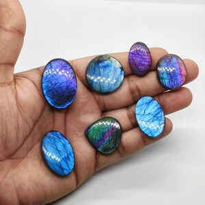 May include: A collection of seven polished labradorite gemstones in various shapes and sizes. The stones display iridescent flashes of blue, purple, and green. The gemstones are held in a hand against a white background, showcasing their natural beauty and color variations.