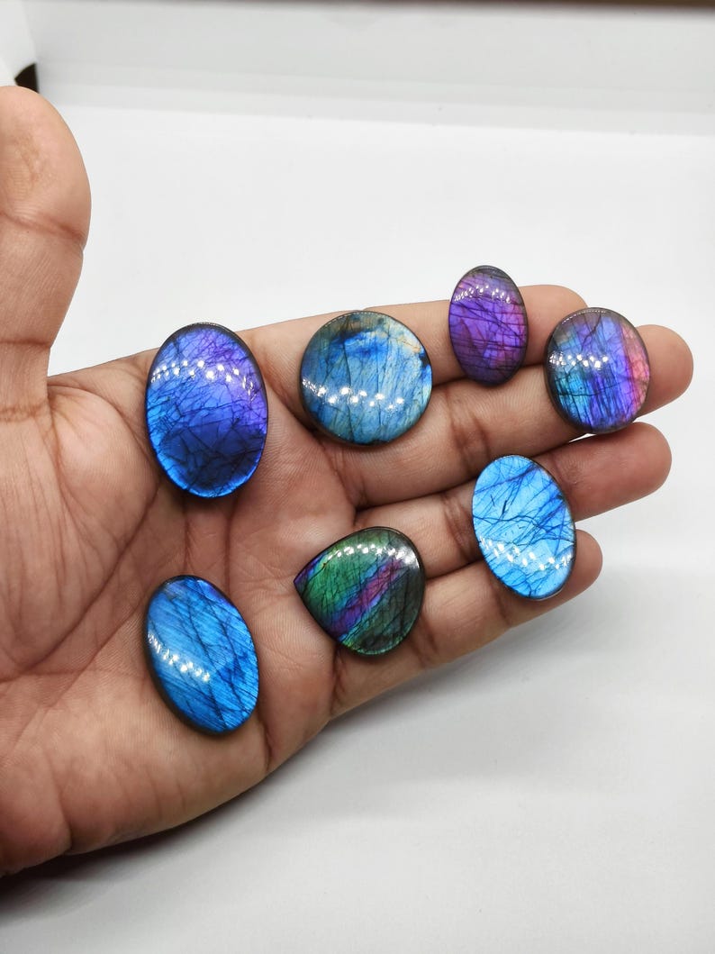 May include: A collection of seven polished labradorite gemstones in various shapes and sizes. The stones display iridescent flashes of blue, purple, and green. The gemstones are held in a hand against a white background, showcasing their natural beauty and color variations.