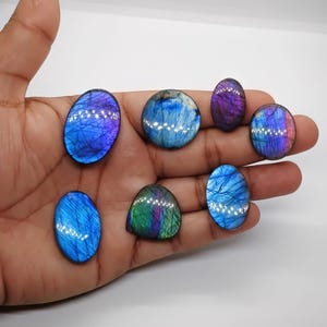 May include: A collection of polished labradorite gemstones in various shapes, including oval and round. The stones display iridescent flashes of blue, purple, and green. The gemstones are held in a hand against a white background.