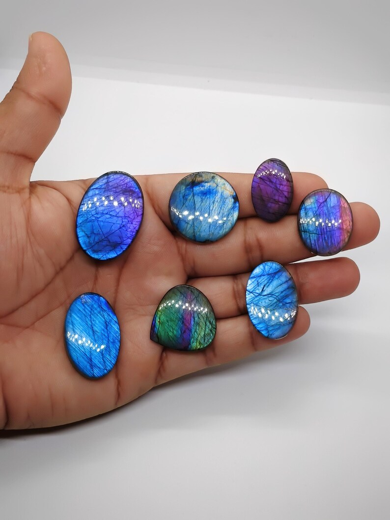 May include: A collection of polished labradorite gemstones in various shapes, including oval and round. The stones display iridescent flashes of blue, purple, and green. The gemstones are held in a hand against a white background.