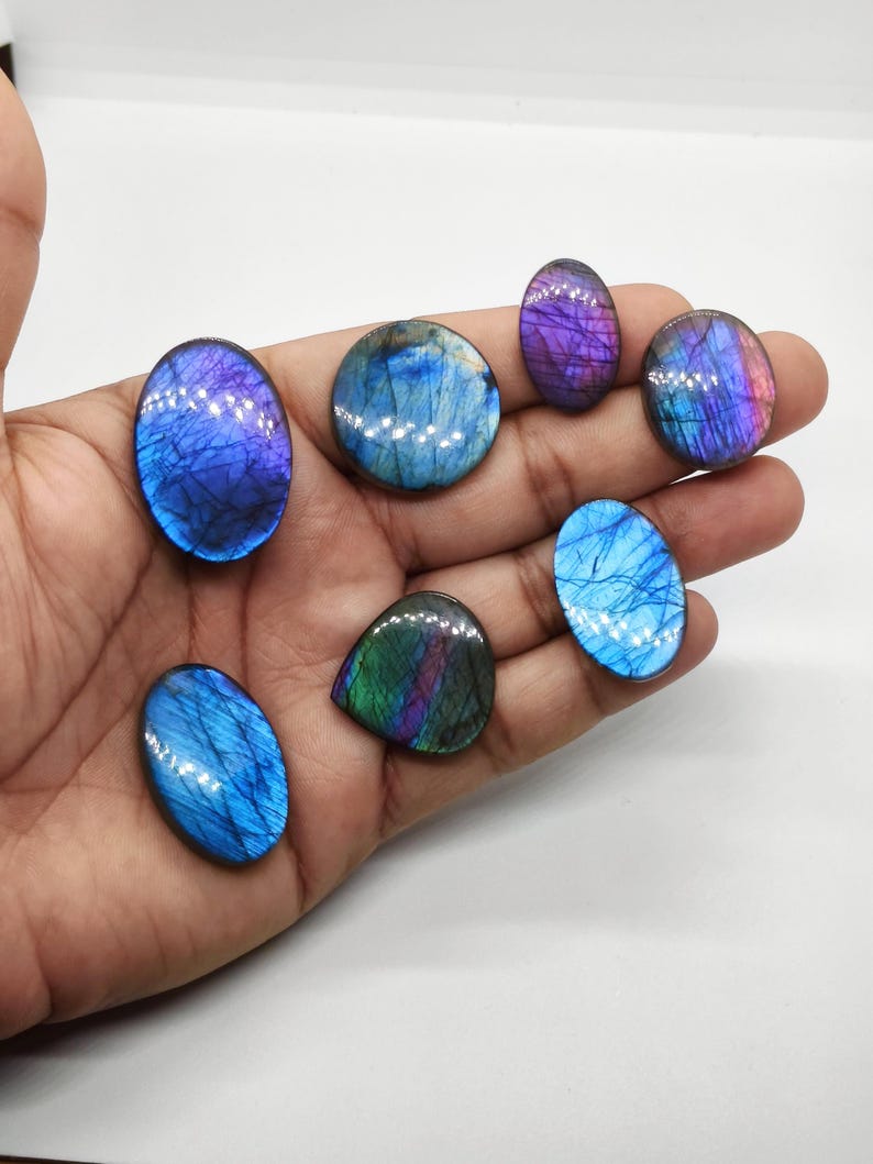 May include: A collection of polished labradorite gemstones in various shapes, including oval and teardrop. The stones display iridescent flashes of blue, purple, and green, with a glossy finish. The gemstones are held in a hand against a white background.