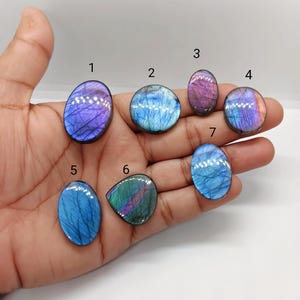 May include: A collection of seven polished labradorite gemstones in various shapes, including oval and teardrop. The stones display iridescent flashes of blue, purple, and green. The gemstones are held in a hand against a white background, showcasing their natural beauty.