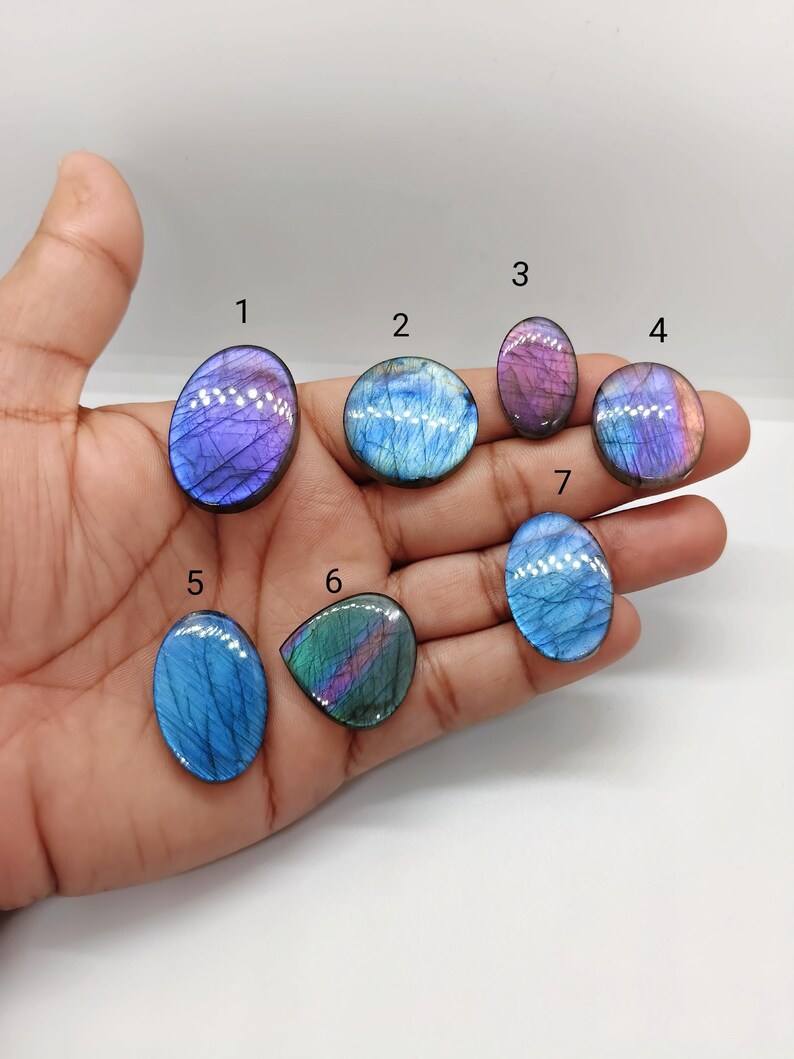 May include: A collection of seven polished labradorite gemstones in various shapes, including oval and teardrop. The stones display iridescent flashes of blue, purple, and green. The gemstones are held in a hand against a white background, showcasing their natural beauty.