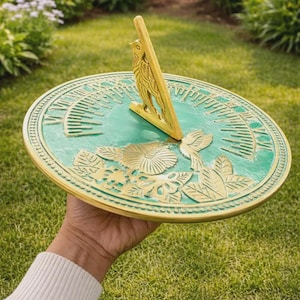Könnte beinhalten: Eine grüne und goldene Sonnenuhr mit Blumen- und Sonnenstrahlendesigns. Die Sonnenuhr wird in einer Hand gehalten und vor dem Hintergrund von grünem Gras und Laub platziert. Der Gnomon ist goldfarben und hat die Form eines Vogels.