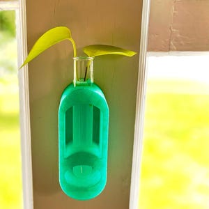 Peut inclure: Un support de propagation mural vert vif avec un tube en verre transparent pour une bouture de plante. Deux feuilles jaune-vert vif dépassent du haut. Le support est monté sur une surface marron clair, avec un arrière-plan vert flou.