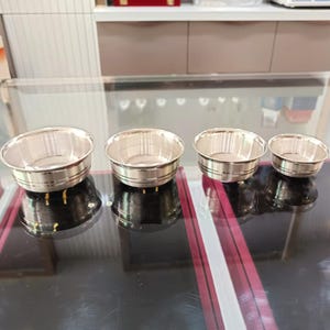 May include: Four small, shiny silver bowls of varying sizes are arranged in a row. Each bowl has a rounded shape with a slightly flared rim and a decorative band near the base. The bowls are displayed on a reflective surface.