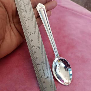 May include: A silver spoon with an ornate handle, approximately 13 cm long, next to a ruler. The spoon has a reflective surface and a decorative design at the handle's end. The background is a solid pink color.