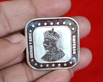 May include: A silver square coin featuring a profile of a crowned figure, with the text "GEORGE V KING" and "EMPEROR" around the edges. The coin has a border with stars.