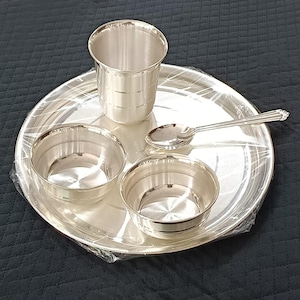 May include: A silver dining set comprising a large round plate, a tall glass, two small bowls, and a spoon. The set is made of shiny metal and is arranged on a dark surface. The items are wrapped in clear plastic.