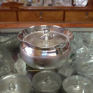 May include: A small, silver-colored bowl with a lid. The bowl has a rounded body and a scalloped rim. A small, round knob sits atop the lid. A leaf design is etched on the side of the bowl.