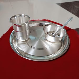 Puede incluir: Un juego de comedor plateado que incluye un plato, una taza, un cuenco pequeño y una cuchara. El plato tiene círculos concéntricos y un diseño central. El juego está hecho de metal plateado brillante y se muestra sobre una superficie roja.