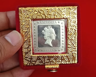 May include: A silver coin featuring Queen Victoria in a gold-colored frame with a red velvet interior. The coin is square and has the words "VICTORIA" and "QUEEN" inscribed. The frame has a decorative pattern.