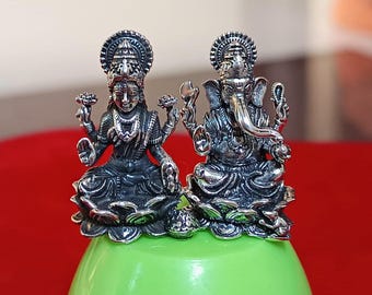 Op de afbeelding: Twee zilverkleurige beelden van de hindoeïstische goden Lakshmi en Ganesha worden getoond. Lakshmi zit en houdt lotusbloemen vast, en Ganesha, met een olifantenkop, zit ook. De beelden staan op een groen oppervlak tegen een rode achtergrond.