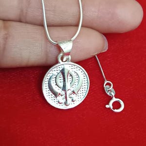 May include: A silver pendant necklace featuring a circular pendant with a detailed religious symbol. The pendant hangs from a delicate silver chain. The background is a solid red color.