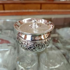 May include: A small, silver-colored container with a lid. The container has a detailed, embossed floral pattern around the base. The lid has a small knob on top. The container is likely used for storing small items.