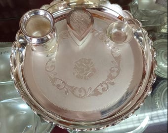 May include: A silver-colored puja thali with a scalloped edge. The thali has a central floral design and includes a small cup, a container, and a heart-shaped holder. The thali is likely used for religious ceremonies.