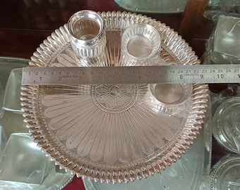May include: A silver-colored metal puja plate with a scalloped edge, featuring three small bowls and a tall glass. A ruler shows the plate is approximately 9 inches in diameter. The plate has a detailed, embossed floral design.