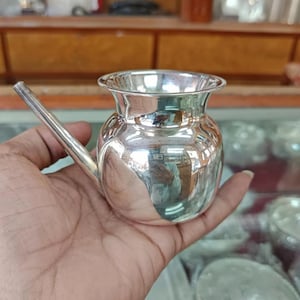 May include: A small, shiny silver vessel with a spout, held in a hand. The vessel has a rounded body and a flared rim. The spout extends from the side. The surface reflects the surrounding environment.