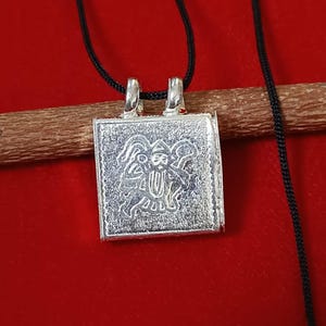 May include: A square silver pendant with an engraved design of a deity, suspended on a black cord. The pendant is detailed with a textured surface and two small loops at the top for the cord. The background is a solid red.