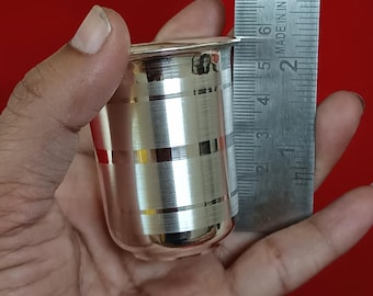 Puede incluir: Una pequeña taza plateada brillante con bandas horizontales. La taza mide aproximadamente 5 cm de alto. La taza se sostiene en una mano sobre un fondo rojo. La taza parece ser de metal.