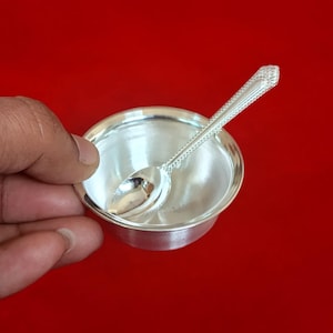 Puede incluir: Un pequeño cuenco plateado brillante con una cuchara a juego. La cuchara descansa dentro del cuenco, que se sostiene sobre un fondo rojo. La cuchara tiene un mango decorativo.