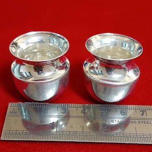 May include: Two small, shiny silver pots with a rounded body and a wide, flared rim. The pots are side-by-side on a red surface, with a stainless steel ruler in the foreground. The pots are approximately 2 inches in height.