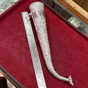 May include: An elaborately decorated silver dagger sheath with floral designs. The curved sheath has an animal head at the end. A ruler is placed next to the sheath, showing measurements in centimetres.