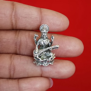 May include: A small, silver-colored statue of a deity playing a stringed instrument, seated on a lotus flower. The statue has intricate details and is set against a red background.