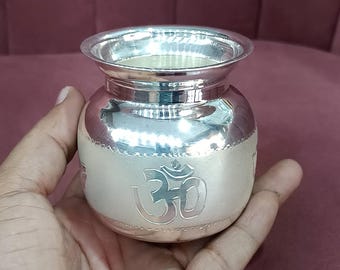 May include: A small, silver-colored pot with a wide rim and a rounded body. The pot features an embossed Om symbol on its side. The pot is held in a hand against a maroon background.