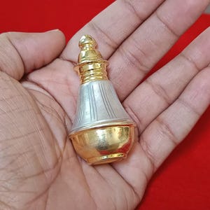 May include: A small, ornate perfume bottle with a gold base and top, and a silver body. The bottle has a bulbous shape and is held in a person's hand. The background is a solid red colour.
