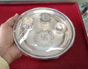 May include: A silver puja plate with three small containers and a central embossed design. The plate is round and has a decorative border. The plate is wrapped in clear plastic and is held by a person.