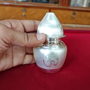 May include: A small, silver-colored pot with a conical lid. The pot has an embossed Om symbol on the front. The pot is held in a person's hand. The background is a red surface and a wooden cabinet.
