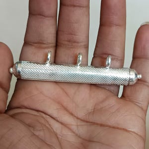 May include: A silver pendant with a cylindrical body and three small loops for hanging. The pendant has a textured, cross-hatched pattern on the main body and rounded end caps. The pendant is held in a hand.
