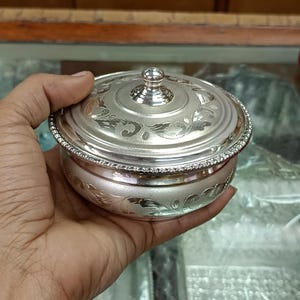 May include: A small, round, silver-colored bowl with a lid, held in a hand. The bowl and lid are adorned with intricate floral patterns and a beaded rim. The lid features a small, decorative knob.