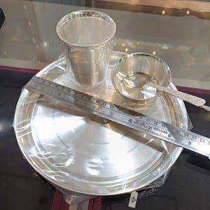Può includere: Un set da pranzo color argento, composto da un piatto, un bicchiere, una piccola ciotola e un cucchiaio. Un righello è posto sul piatto. Il set è avvolto in plastica trasparente. Il piatto misura circa 23 cm di diametro.