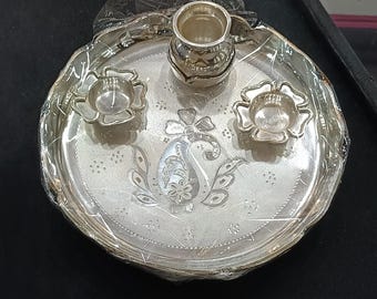 May include: A silver-colored puja thali set, featuring a central plate with floral and paisley engravings. The set includes two small flower-shaped bowls and a small cup. The thali is wrapped in clear plastic.