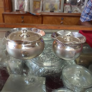 May include: Two silver-colored, lidded containers with a floral design. The containers have a round shape with a ruffled edge on the lid. The lids have a small, round knob on top. The containers are sitting on a glass surface.