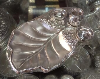 May include: A silver-colored leaf-shaped serving dish with three small, round containers. The dish has a textured surface with detailed leaf veins. The containers are likely for condiments or small portions.